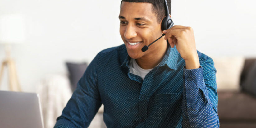 African american young man customer support call center operator or receptionist sitting at the workplace in a modern office consulting a client, uses a headset, smiles friendly